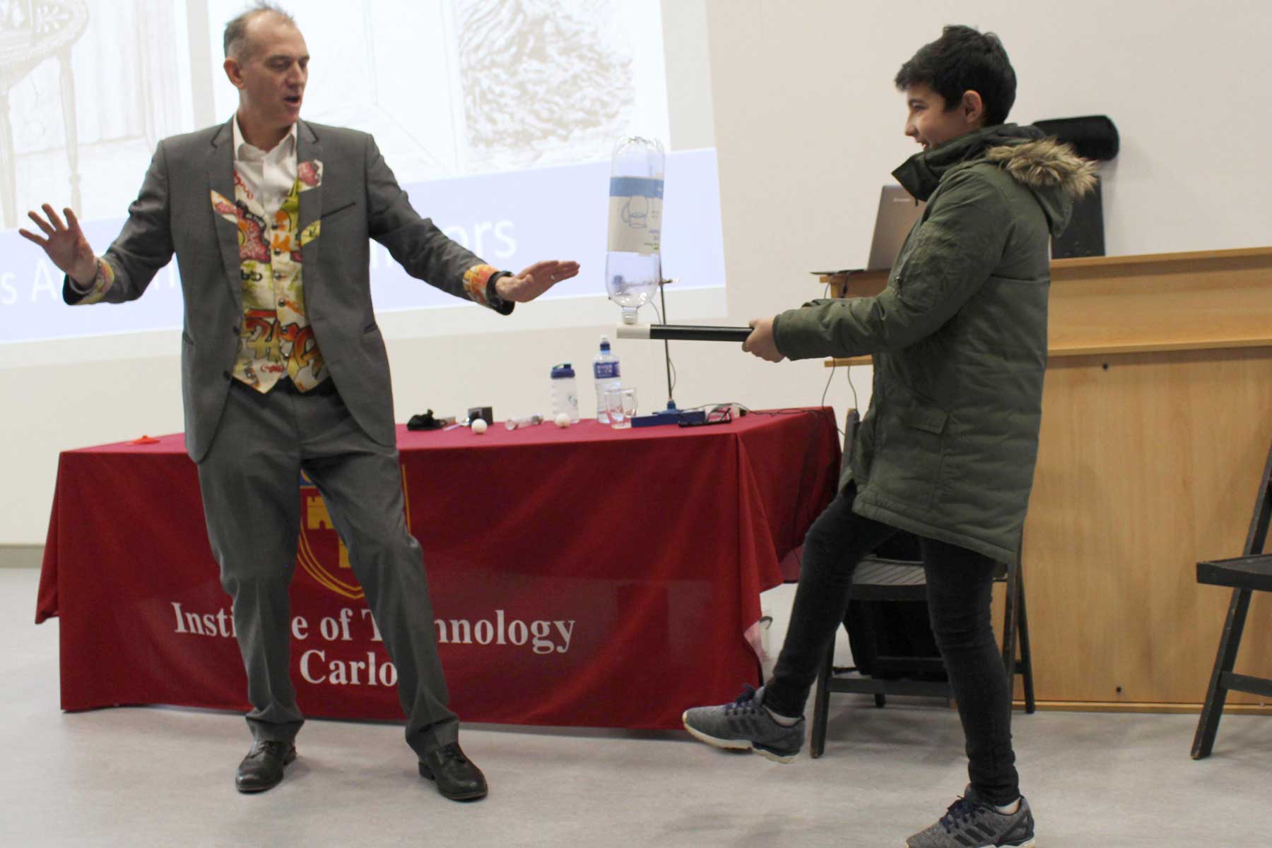 An empty water bottler is balanced on the end of a wand by a volunteer standing on one leg as Dr Ken says 'Abracadabra'
